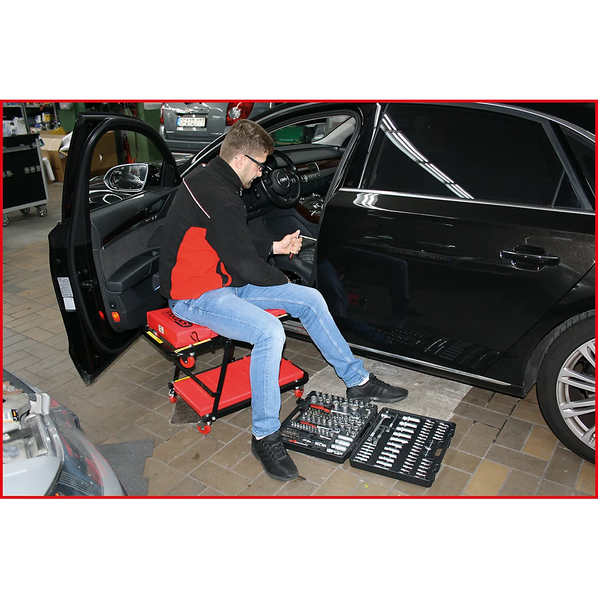 Un homme dans un atelier répare une voiture. Il est assis sur un tabouret rouge et a des boîtes à outils devant lui.